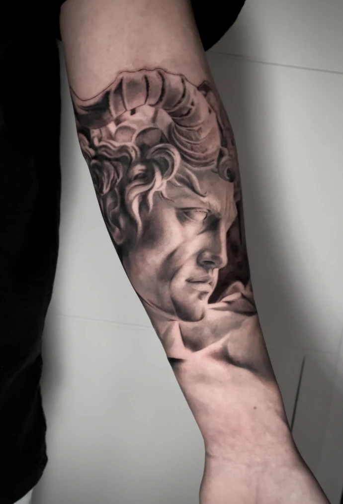 Dieses beeindruckende Black-and-Grey Realismus-Tattoo am Unterarm zeigt ein kraftvolles Porträt eines antiken Fauns, das durch seine meisterhafte Detailtiefe und plastische Schattierung besticht. Die kunstvolle Umsetzung im Realismus-Stil fängt die markanten Gesichtszüge und die charakteristischen Hörner mit einer außergewöhnlichen Dreidimensionalität ein, wodurch ein zeitloses, mythologisches Kunstwerk direkt auf der Haut entsteht