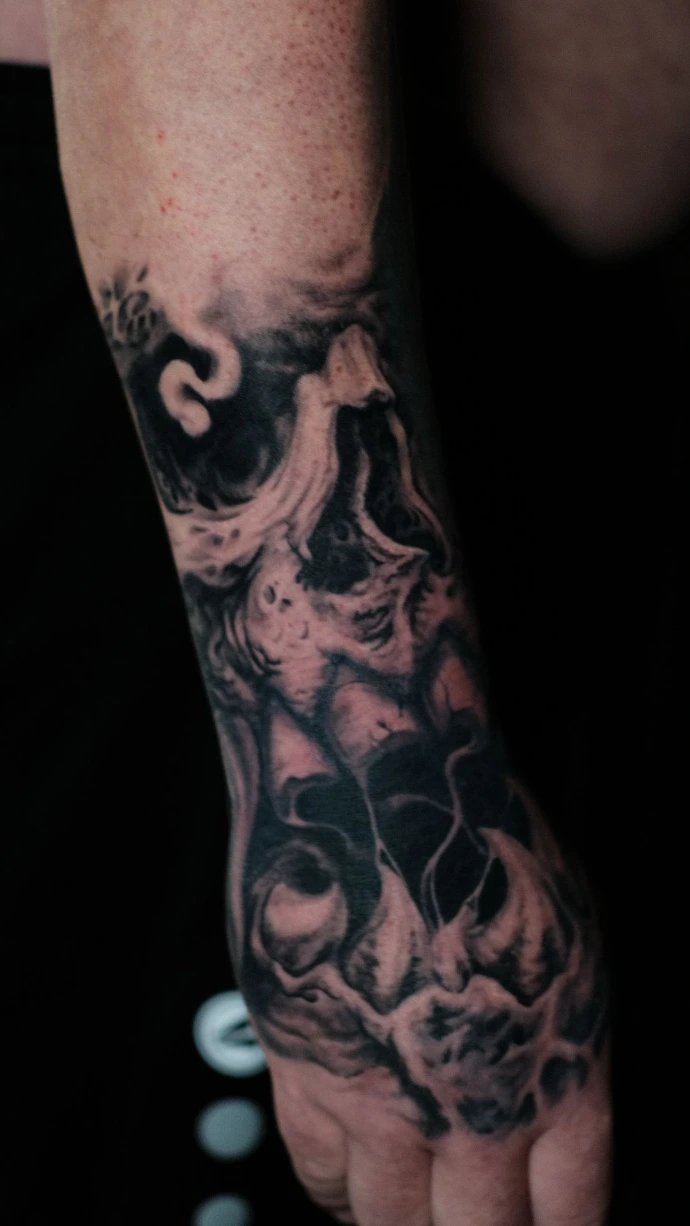 Dieses düstere Black-and-Grey Realismus-Tattoo auf dem Handrücken zeigt eine abstrakte, fließende Interpretation eines Totenkopf-Motivs, das nahtlos in den Unterarm übergeht. Die meisterhafte Umsetzung im Dark-Art-Stil besticht durch ihre plastische Tiefe und die organischen, fast rauchähnlichen Strukturen, die durch feinste Schattierungen und harte Kontraste eine beeindruckende, unheimliche Dynamik direkt auf der Hand erzeugen.