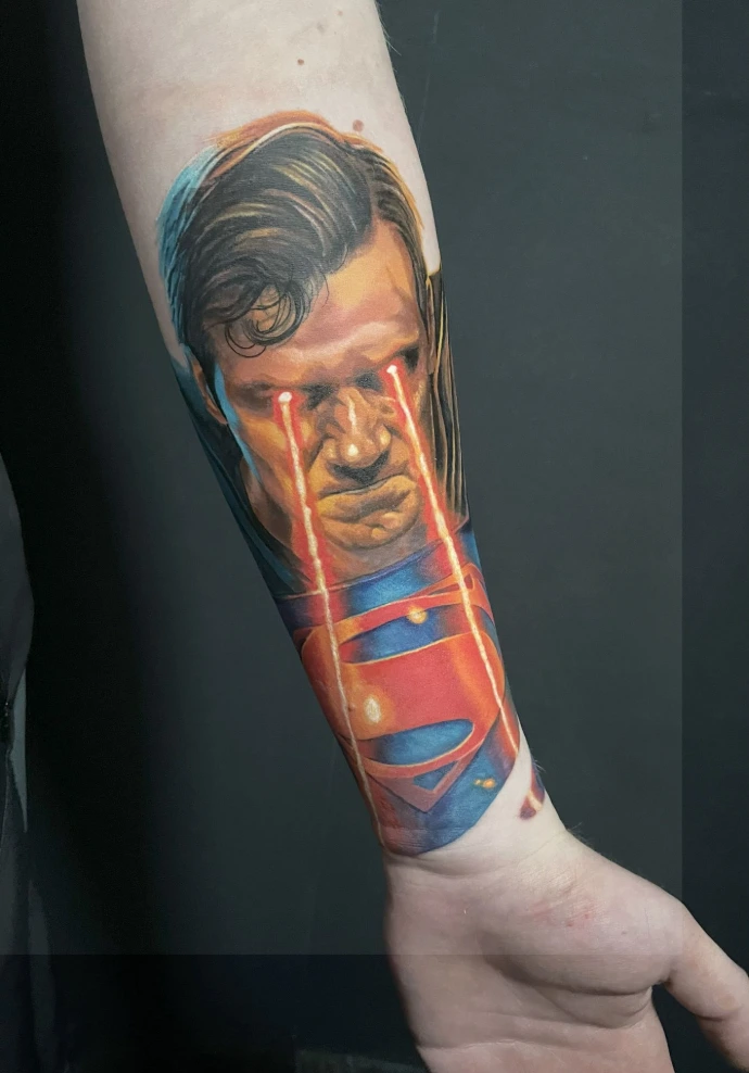 Dieses kraftvolle Realismus-Tattoo am Unterarm zeigt ein dynamisches Porträt von Superman mit leuchtendem Hitzeblick, das durch seine außergewöhnliche Farbtiefe und Lichteffekte besticht. Die meisterhafte Umsetzung im Farbrealismus nutzt intensive Kontraste zwischen den kühlen Blautönen des Anzugs и den glühend roten Laserstrahlen, um eine beeindruckende Dreidimensionalität und Energie direkt auf der Haut zu erzeugen