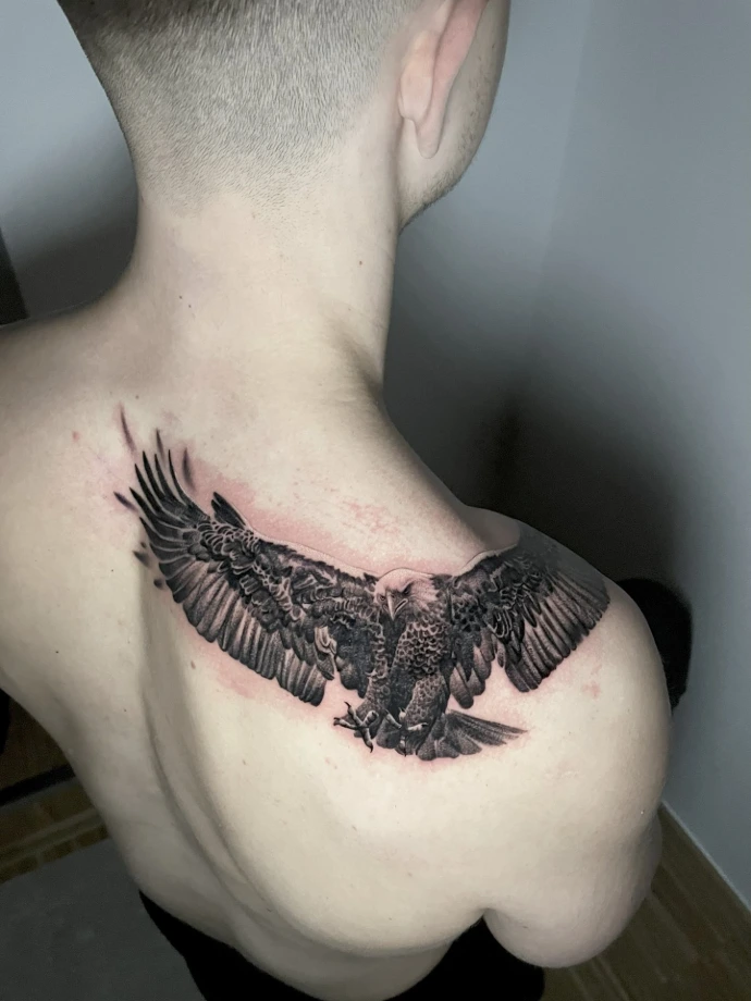 Dieses beeindruckende Black-and-Grey Realismus-Tattoo auf dem Rücken zeigt einen majestätischen Adler im Sturzflug, der durch seine präzise Detailtiefe und kraftvolle Dynamik besticht. Die meisterhafte Umsetzung im Realismus-Stil nutzt feinste Schattierungen, um die Textur jeder einzelnen Feder plastisch hervorzuheben, während die anatomisch angepasste Platzierung über Schulterblatt und Nacken die imposante Flügelspannweite perfekt in Szene setzt.