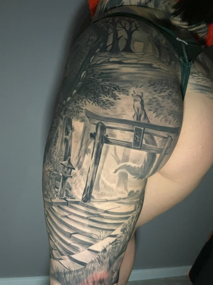 Dieses exzellente Black-and-Grey Realismus-Tattoo auf der Gesäßpartie beeindruckt durch seine architektonische Tiefe und detailreiche Ausarbeitung japanischer Mythologie. Im Zentrum steht eine perspektivisch perfekt gesetzte Treppe, die zu einem imposanten Torii-Tor führt, auf dem eine stolze Kitsune (Fuchsdämon) wacht. Die meisterhafte Umsetzung im Realismus-Stil nutzt feinste Schattierungen, um die steinerne Textur der Stufen und die feine Fellstruktur des Fuches plastisch hervorzuheben, wodurch das großflächige Motiv harmonisch mit der weiblichen Anatomie verschmilzt