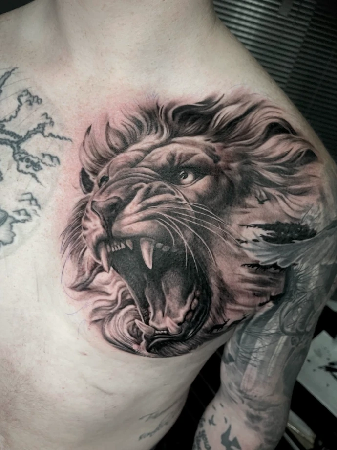 Dieses beeindruckende Realismus-Tattoo auf der Brust zeigt ein kraftvolles Porträt eines brüllenden Löwen, das durch seine präzise Detailtiefe und lebendige Dynamik besticht. Die kunstvolle Umsetzung in Black-and-Grey Realismus nutzt feinste Schattierungen, um die Textur der Mähne und den intensiven Ausdruck des Raubtieres plastisch hervorzuheben, wodurch ein majestätisches und zeitloses Motiv auf der Haut entsteht