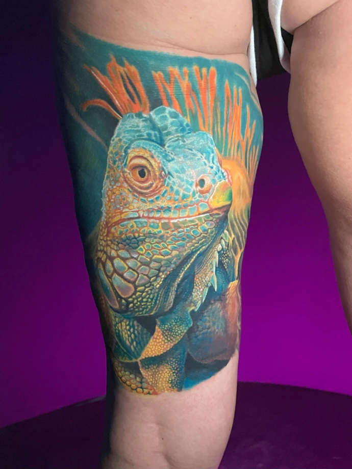 Dieses kraftvolle Realismus-Tattoo am Oberschenkel zeigt ein detailreiches Porträt einer Iguana, das durch seine lebendige Farbtiefe und präzise Texturen der Schuppen besticht. Die kunstvolle Umsetzung im Farbrealismus kombiniert leuchtende Türkis- und Orangetöne, um eine plastische, naturnahe Optik zu erzeugen, die das exotische Motiv auf der Haut perfekt in Szene setzt.