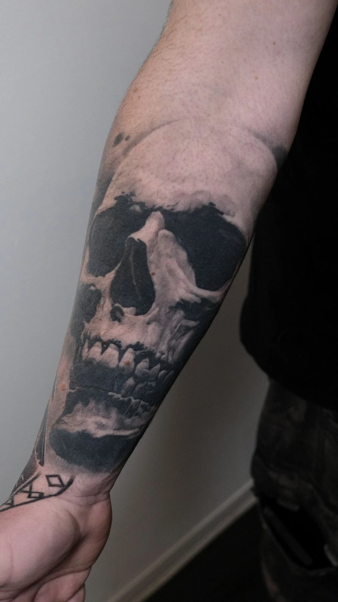 Dieses ausdrucksstarke Black-and-Grey Realismus-Tattoo am Unterarm zeigt ein detailreiches Totenkopf-Motiv, das durch seine intensiven Kontraste und plastische Tiefe besticht. Die meisterhafte Schattierung und präzise Ausarbeitung der Knochenstruktur verleihen dem Skull-Tattoo eine düstere, kraftvolle Optik, die sich perfekt an die Anatomie des Arms anpasst und als zeitloses Kunstwerk in Graustufen überzeugt.