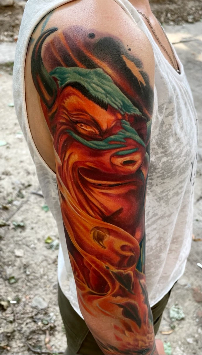 Dieses feurige Realismus-Tattoo am Oberarm zeigt ein intensives Porträt eines lachenden Dämons, das durch seine lebendige Farbtiefe und dynamische Komposition besticht. Die meisterhafte Umsetzung im Farbrealismus nutzt eine glühende Palette aus Orange-, Rot- und Gelbtönen, die durch kontrastreiches Schwarz und kühle türkisfarbene Akzente perfekt ergänzt werden, um eine plastische, fast greifbare Hitze auf der Haut zu erzeugen.