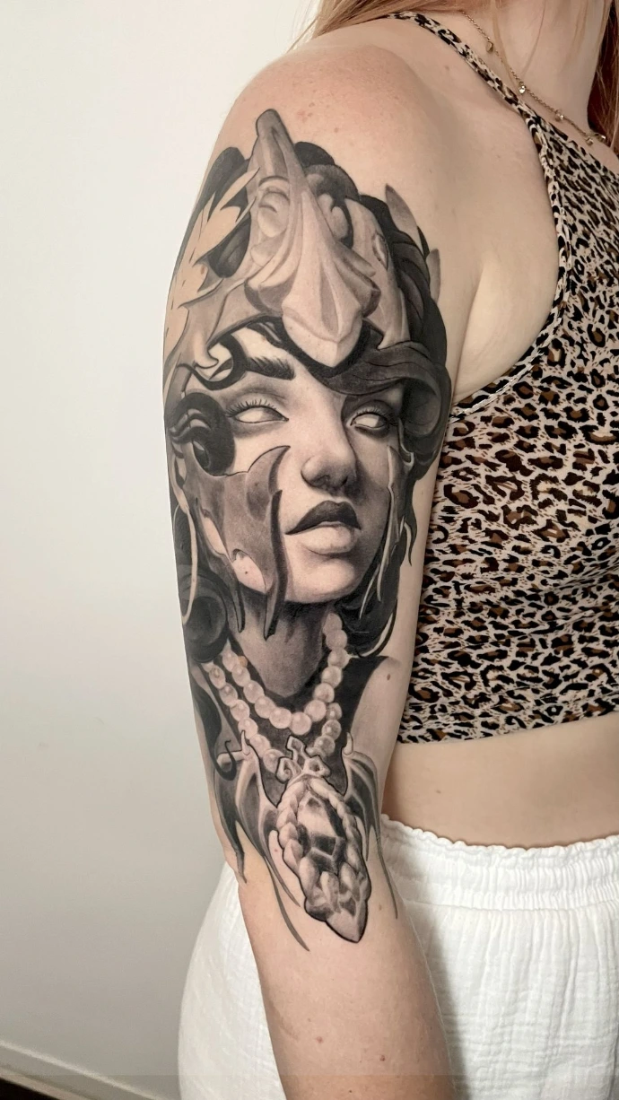 Dieses beeindruckende Black-and-Grey Neo-Traditional Tattoo am Oberarm zeigt ein mystisches Frauenporträt mit einer markanten Knochenmaske und filigranem Perlenschmuck. Die Arbeit besticht durch die für den Neo-Trad-Stil typischen kräftigen Outlines und die meisterhafte Schattierung, die dem Gesicht und den organischen Elementen eine außergewöhnliche Tiefe verleiht. Die Kombination aus düsterer Symbolik und eleganter Linienführung macht dieses großflächige Motiv zu einem ausdrucksstarken Kunstwerk auf der Haut.
