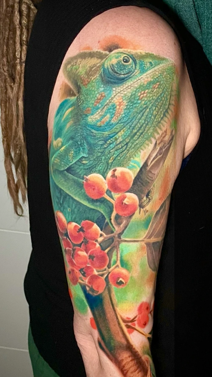 Dieses beeindruckende Realismus-Tattoo am Oberarm zeigt ein detailreiches Chamäleon auf einem Ast mit roten Beeren, das durch seine lebendige Farbtiefe und präzise Texturen besticht. Die kunstvolle Umsetzung im Farbrealismus nutzt kräftige Grün- und Orangetöne, um eine plastische, naturnahe Optik zu erzeugen, die das Motiv auf der Haut zum Leben erweckt