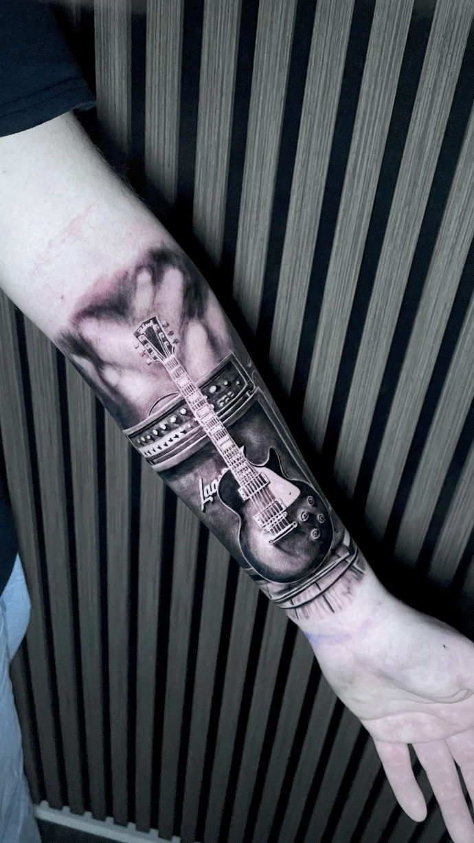 Dieses detailreiche Black-and-Grey Realismus-Tattoo am Unterarm setzt eine klassische Gibson Les Paul E-Gitarre vor einem Marshall-Verstärker perfekt in Szene. Die meisterhafte Umsetzung im Realismus-Stil besticht durch die präzise Ausarbeitung glänzender Oberflächen, feinster Saiten und technischer Details, während die rauchigen Schattierungen im Hintergrund eine rockige Atmosphäre und beeindruckende Tiefe auf der Haut erzeugen.