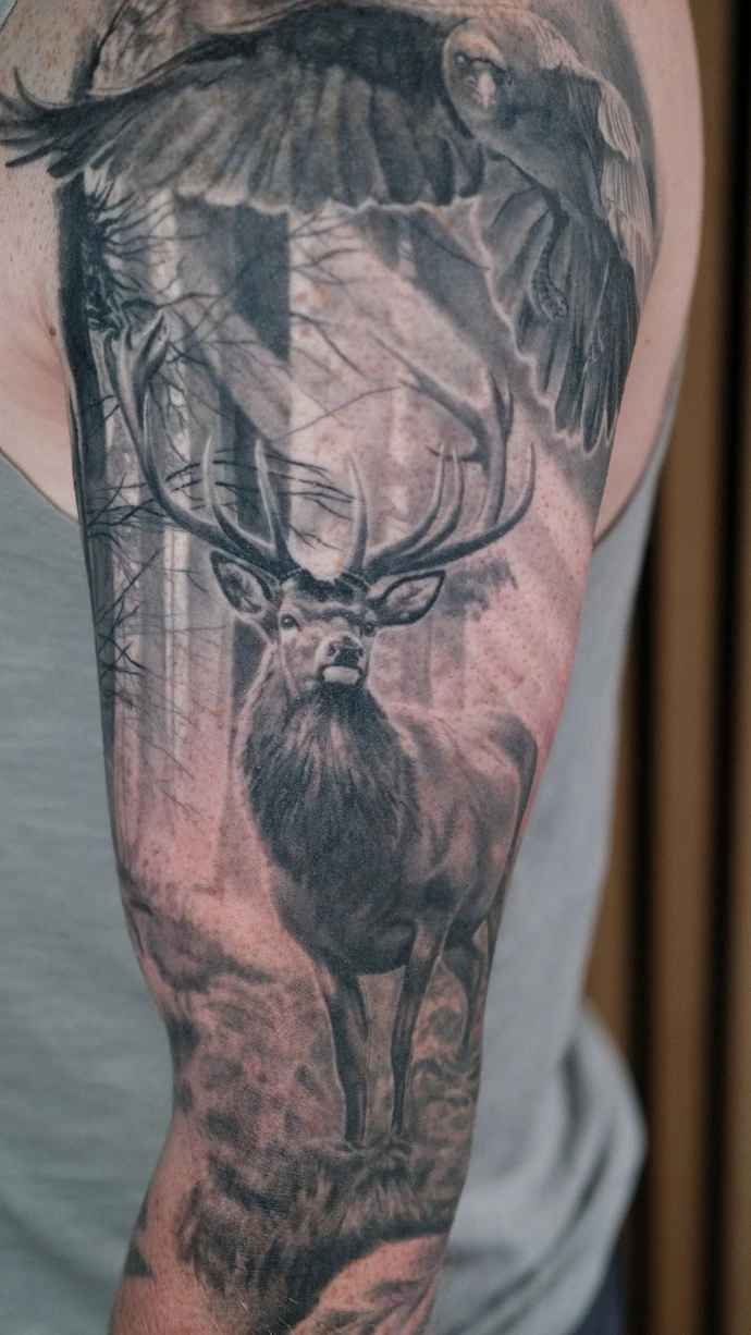 Dieses stimmungsvolle Black-and-Grey Realismus-Tattoo am Oberarm zeigt eine majestätische Waldszene mit einem Hirsch und einem fliegenden Raben im Hintergrund. Die meisterhafte Umsetzung im Realismus-Stil besticht durch die detailreiche Ausarbeitung des Hirschgeweihs und der Federstruktur des Raben, während die einfallenden Lichtstrahlen zwischen den Bäumen eine beeindruckende Tiefe und eine mystische Atmosphäre auf der Haut erzeugen.