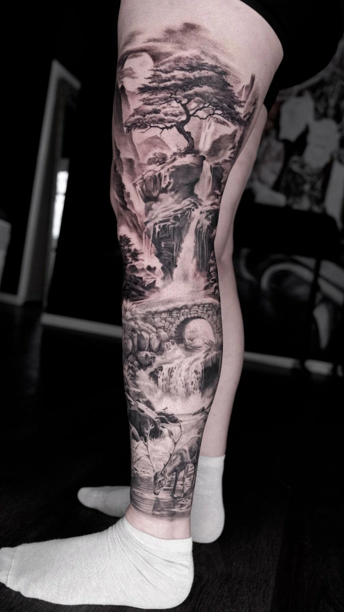 Dieses beeindruckende Full-Leg Tattoo im Black-and-Grey Realismus zeigt eine detailreiche Naturlandschaft, die sich harmonisch über das gesamte Bein erstreckt. Das Motiv kombiniert meisterhaft verschiedene Elemente wie einen majestätischen Baum auf einem Felsen, kaskadierende Wasserfälle, eine klassische Steinbrücke und ein friedlich trinkendes Reh am Wasserlauf. Die präzise Schattierung und perspektivische Tiefe im Realismus-Stil verwandeln die gesamte Beinfläche in ein lebendiges, zusammenhängendes Kunstwerk voller Ruhe und Dynamik.