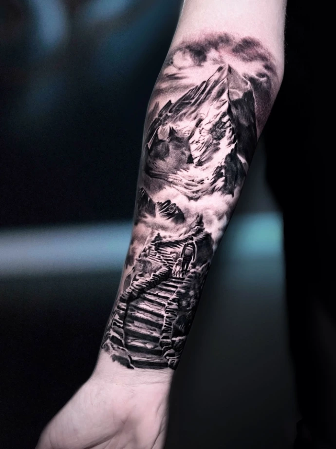 Dieses beeindruckende Black-and-Grey Realismus-Tattoo am Unterarm zeigt eine atmosphärische Berglandschaft mit einer steineren Treppe, die zu einem majestätischen Gipfel führt. Das Motiv besticht durch die detailreiche Darstellung eines Wanderers mit Hund, der den Pfad erklimmt, eingebettet in dramatische Wolkenformationen. Die meisterhafte Umsetzung im Realismus-Stil nutzt starke Kontraste und feinste Schattierungen, um eine außergewöhnliche Tiefe und ein Gefühl von Freiheit und Abenteuer auf der Haut zu erzeugen.